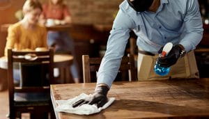 A Waiter with Mask and Gloves Cleaning a Table in a Caffee Using Detergent  